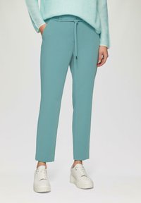 Turquoise joggings met een verstelbare tailleband, zijzakken en een slim fit, gecombineerd met witte platform sneakers. Vlakke textuur.