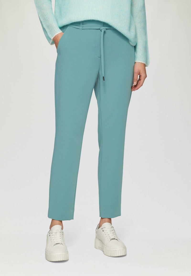 Turquoise joggings met een verstelbare tailleband, zijzakken en een slim fit, gecombineerd met witte platform sneakers. Vlakke textuur.