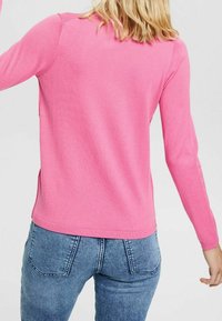 Dopasowany sweter z długim rękawem w jaskrawym różowym kolorze, wykonany z dzianiny, z prążkowanymi mankietami i krawędzią. W połączeniu z jasnoniebieskimi dżinsami.