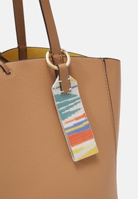 Brun læder tote bag har en glat tekstur, en ring i guldhardware, og et mønstret stofmærke med blå, orange og gule striber.