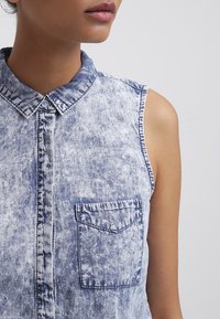 Gros plan d'une personne portant une chemise en jean sans manches avec un col et une poche poitrine, sur un fond clair uni.