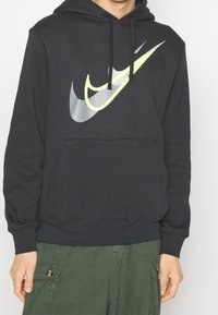 Sudadera con capucha negra que presenta un gran logo de Nike multicolor en la parte delantera, con acentos en gris, amarillo y negro. Bolsillo tipo canguro en la parte frontal.