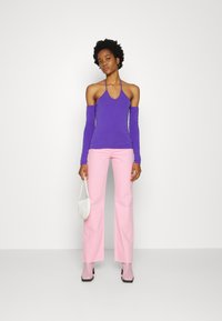Top roxo com alças e mangas longas, combinado com calças flare cor-de-rosa claro. Modelo segura uma pequena mala branca e usa botas de salto cor-de-rosa pálido.