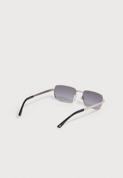 Lunettes de soleil rectangulaires à monture argentée avec verres gris foncé et embouts noirs sur les branches, posées sur une surface blanche.