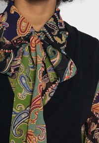 Wielokolorowy szal w wzór paisley z zielonymi, czarnymi i niebieskimi akcentami, związany w węzeł na szyi, na czarnym, teksturowanym swetrze.