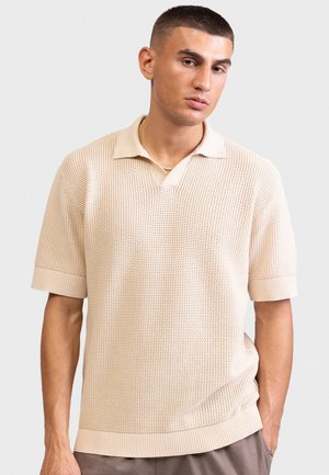 Νεαρός άνδρας με κοντά σκούρα μαλλιά που φοράει ανοιχτό μπεζ πόλο πουλόβερ με υφή και καφέ παντελόνια μπροστά από ένα απλό φόντο.