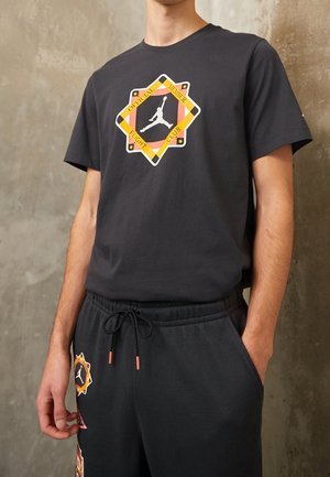 Czarny bawełniany t-shirt z kolorową grafiką sylwetki koszykarza w kształcie diamentu. W połączeniu z luźnymi czarnymi spodniami dresowymi z podobnymi grafikami.