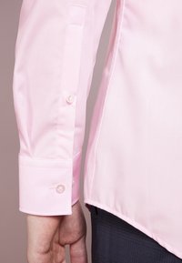 Lichtroze shirtmouw met een gladde textuur, voorzien van een enkele knoopsluiting en een gebogen zoom. Subtiele glans zichtbaar in de stof.