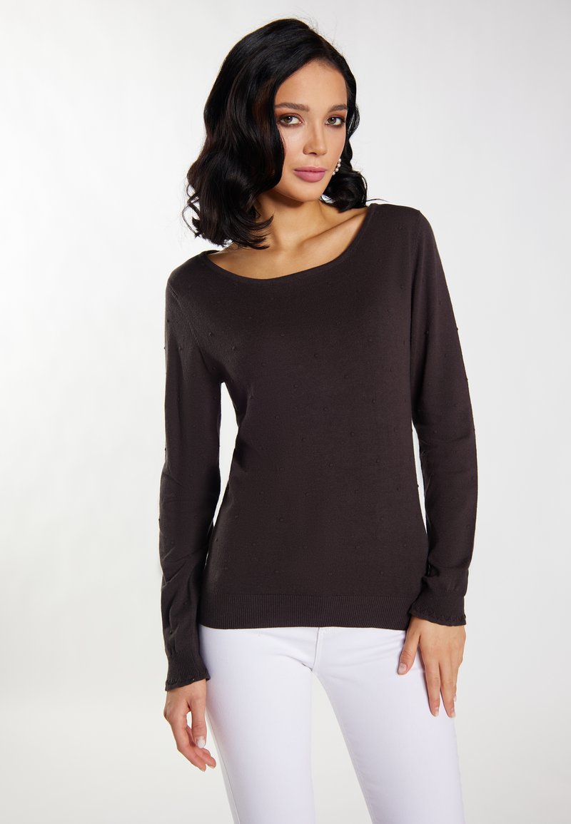 Dunkelbrauner Pullover mit langen Ärmeln und weitem Ausschnitt sowie strukturierten Punkten. Kombiniert mit weißen Hosen, die eine figurbetonte Silhouette zeigen.