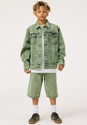 Grön denimjacka med knappar, två framsidorfickor, kombinerad med matchande gröna denimshorts. Vit skjorta under och bruna sneakers.