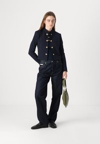 Giacca cropped blu navy con bottoni dorati, abbinata a jeans scuri a vita alta. Modello che tiene una borsa verde foresta.