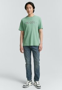 Camiseta de algodón verde claro con un estampado de logo en la parte frontal, combinada con jeans azules y zapatillas en blanco y negro. Corte casual, mangas cortas.