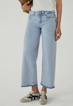 Osoba nosząca jasnoniebieskie szerokie, krótkie spodnie jeansowe, białe trampki z czarnymi paskami oraz biały top i trzymająca brązową kurtkę.