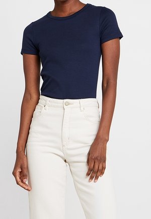 Person trägt ein figurbetontes, marineblaues kurzärmeliges Hemd, das in hochtaillierte weiße Jeans mit vorderen Taschen und Knopfverschluss gesteckt ist.