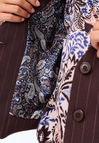 Brązowa kurtka z prążkowanym wzorem, z wzorzystą niebieską podszewką w paisley. Zawiera brązowy guzik i floralne akcenty.