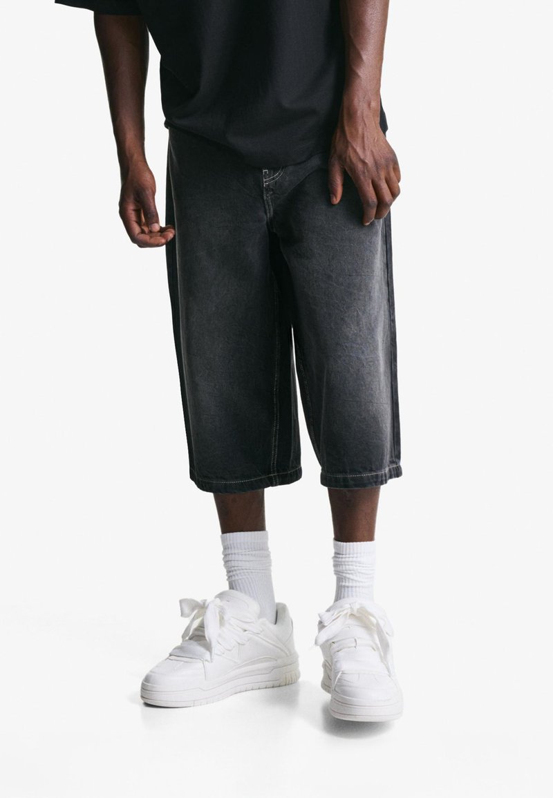 Man wearing black oversized shorts, white crew socks, and white sneakers, standing against a plain white background.