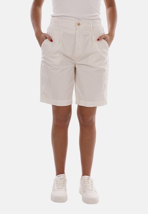 Personne portant un short beige jusqu'aux genoux avec des poches devant, un haut blanc et des baskets blanches, debout les mains dans les poches.