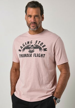 Mężczyzna z krótkimi ciemnymi włosami i brodą, ubrany w jasnoróżowy t-shirt z czarnym wizerunkiem orła i napisem "Raging Storm Thunder Flight", ręce w kieszeniach.