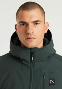 Chaqueta aislante de color verde oscuro con collar alto, cierre frontal con cremallera y parche de logo. Presenta un acabado texturizado y un bolsillo lateral con cremallera.