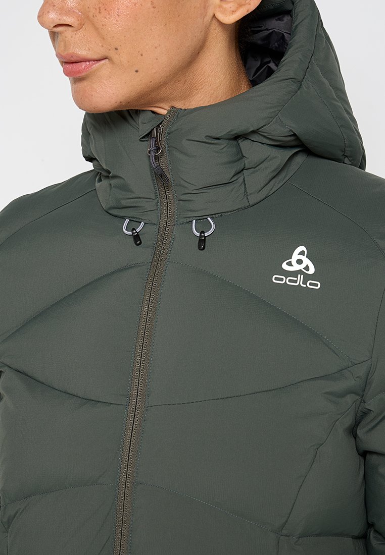 Frau in einer grünen Odlo gepolsterten Jacke mit hohem Kragen und Reißverschluss, die das obere Torso und das Gesicht im Profil zeigt.