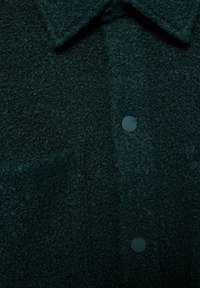 Camicia verde scuro con bottoni, dalla texture strutturata, con colletto, realizzata in tessuto morbido e peloso, con bottoni piatti. Vista ravvicinata che evidenzia i dettagli del materiale.