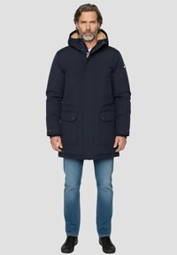 Veste longue marine avec capuche doublée de fourrure, fermeture éclair à l'avant et boutons-pression. Comprend deux poches avant et une finition légèrement texturée. Portée sur un jean.