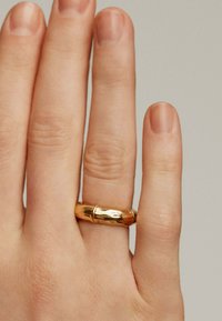 Anillo de oro con una superficie texturizada, diseñado con una forma lisa y redondeada, llevado en el dedo anular de una mano sobre un fondo neutro.