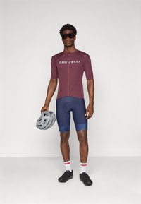 Maillot de cyclisme à manches courtes couleur bordeaux avec fermeture éclair et logo "Castelli", associé à un short bleu marine. Le modèle tient un casque gris et porte des chaussures noires.