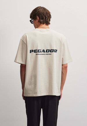 Man draagt een lichtbeige oversized T-shirt met tekst "PEGADOR Certified Authentic Streetwear" op de rug, gecombineerd met een zwarte broek en een zonnebril.