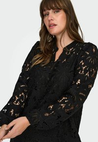 Crna čipkasta majica s cvjetnim izrezanim uzorcima, dugim rukavima s elastičnim manžetama i visokim ovratnikom. Tkanina ima teksturirani, slojeviti izgled.