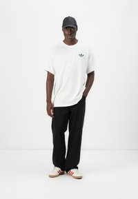 Tricou alb din bumbac cu un logo verde Adidas pe pieptul stâng, asortat cu pantaloni largi negri și adidași albi cu dungi roșii.