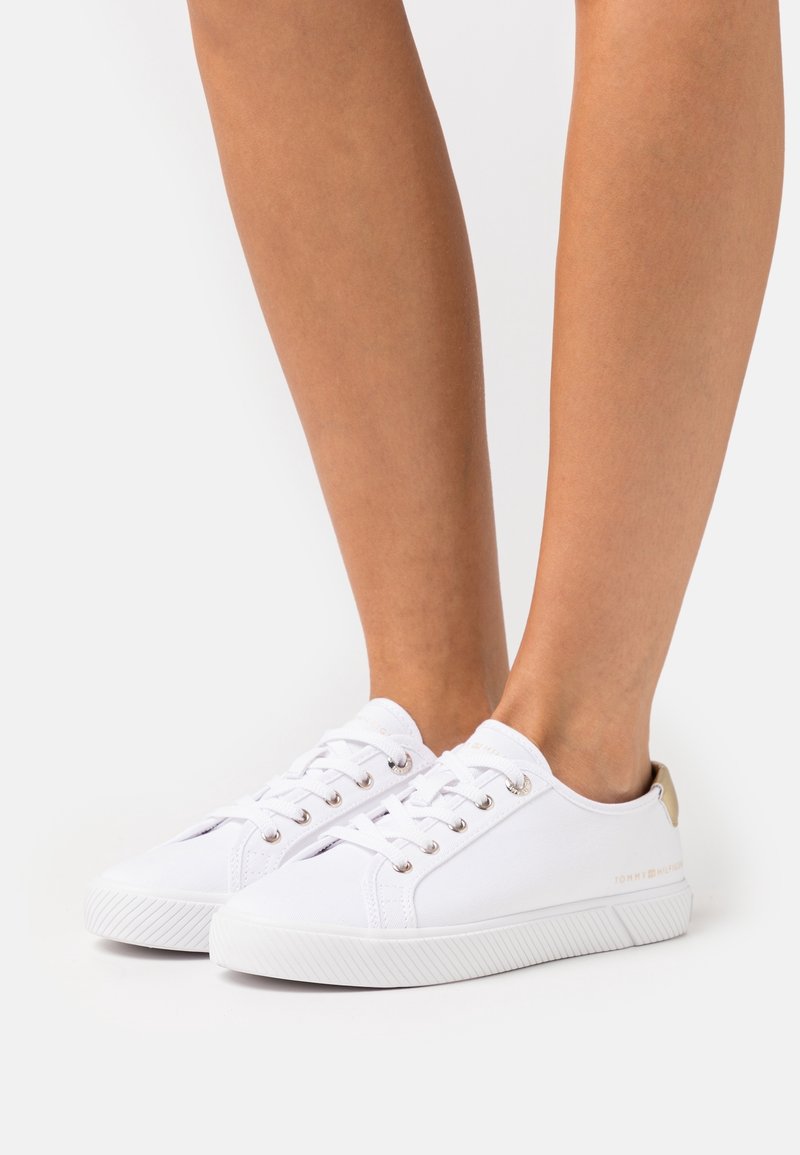 Sneaker bianche in tela con suola texturizzata, dotate di occhielli metallici e un accento dorato sul tallone. Design semplice con lacci piatta.