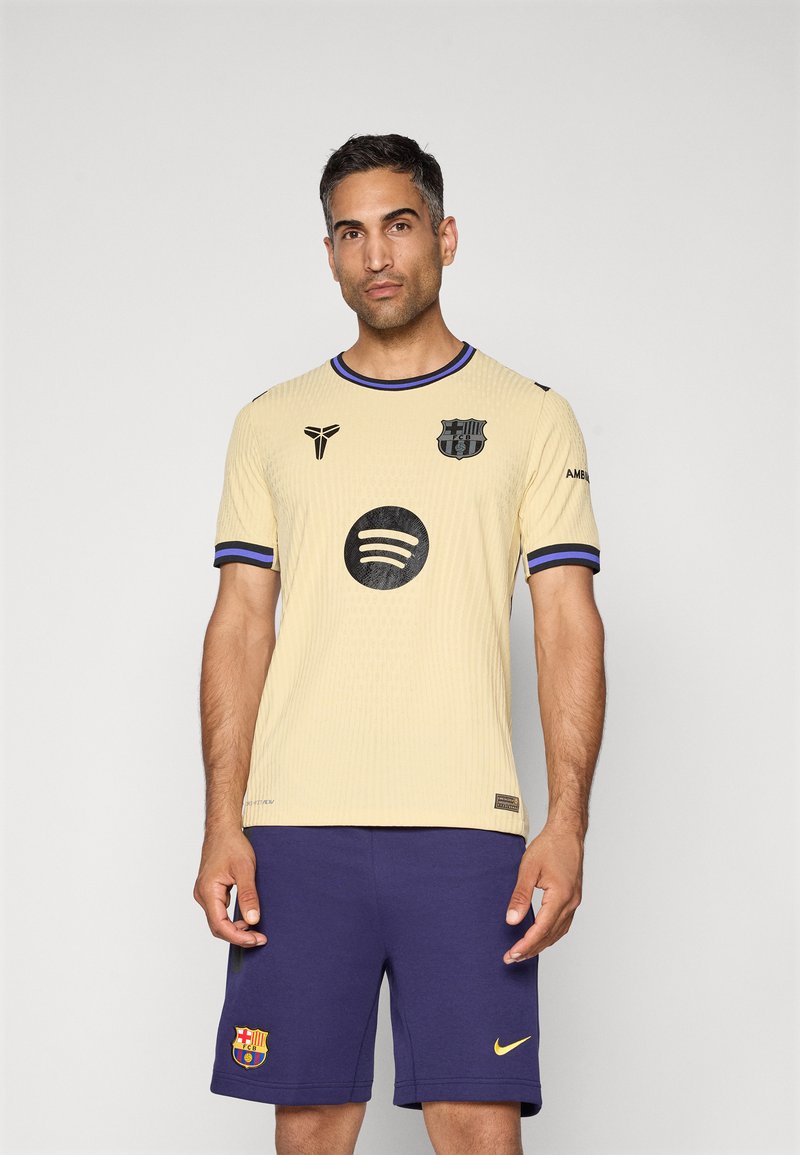 Lysegul trøje med sorte accenter, rund halsudskæring, ribbet tekstur, med et logo, sort Spotify-cirkel og blå ærmeafslutninger. Sammen med marineblå shorts.