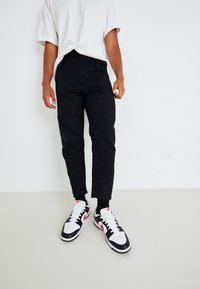 Svarta tapered byxor med en slät textur, tillsammans med vita sneakers med svarta detaljer och röda accenter. Vit oversized t-shirt.