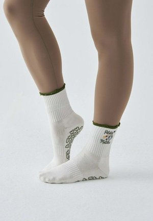 Stopy w białych, antypoślizgowych skarpetkach z zielonym obszyciem i napisem „Pilates & Matcha”, ustawione na palcach na jednolitym tle.