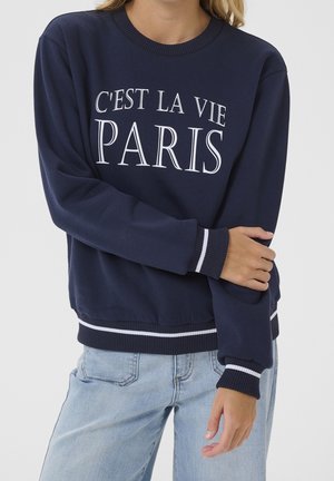 Osoba ubrana w granatową bluzę z białym napisem "C'EST LA VIE PARIS," w zestawieniu z jasnoniebieskimi dżinsami, z założonymi na piersi skrzyżowanymi ramionami.