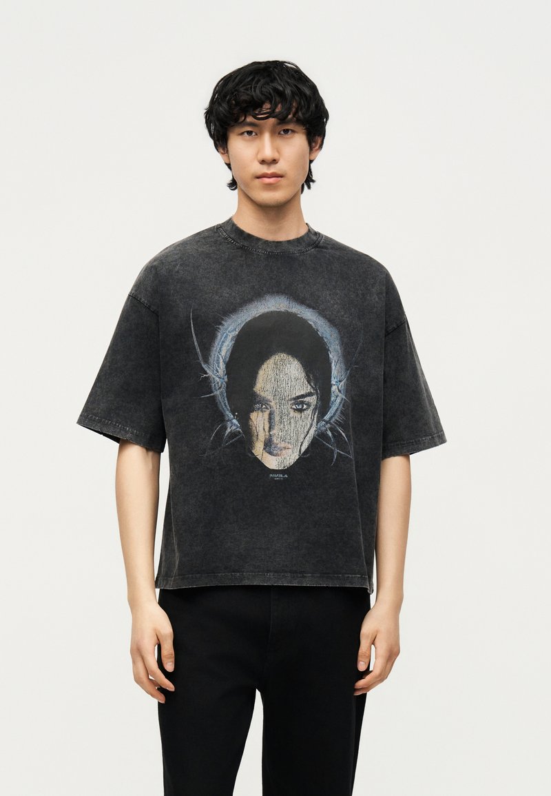 Νέος άνδρας με μαύρα κατσαρά μαλλιά που φοράει μια σκουρογκρί φαρδιά μπλούζα με μεγάλο καλλιτεχνικό γραφικό ενός προσώπου μπροστά.