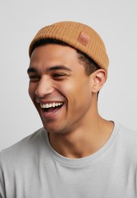 Brown knitted beanie with a ribbed texture and a leather patch. Worn with a light gray crewneck shirt.