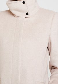 Cappotto di lana beige con colletto alto, chiuso da due bottoni neri e con tasche laterali, mostrato dalla parte del petto fino a metà braccio.