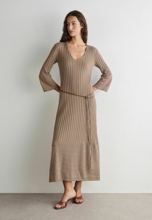 Femme portant une robe midi en maille côtelée taupe avec manches cloche, col en V, ceinture fine et sandales marron ouvertes à talons, debout contre un mur uni.