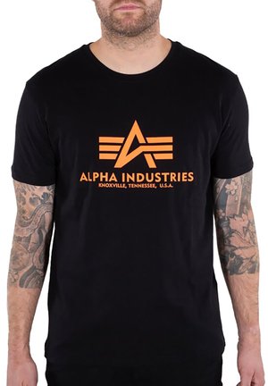 Mann mit Arm-Tattoos trägt ein schwarzes T-Shirt mit dem orangefarbenen Alpha Industries-Logo und dem Text "Knoxville, Tennessee, U.S.A." auf der Vorderseite.