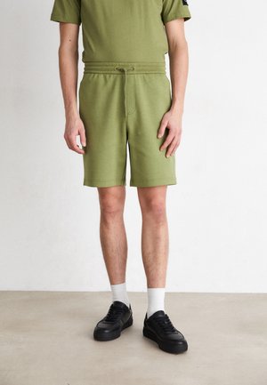 Pantalones cortos de color verde oliva con una cintura ajustable y cordón, fabricados en material suave. Combinados con zapatillas negras y calcetines blancos hasta el tobillo.