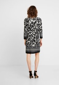 Robe noire et blanche à motif de feuilles, manches longues, et ourlet avec bande décorative. Paire de chaussures à talons hauts noires.