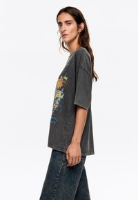 Sivý oversized tričko s krátkymi rukávmi, na prednej strane s farebným grafickým dizajnom. Na sebe má vysoké modré džínsy.