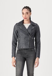 Svart skinnmotorsykkeljakke med polstrede skuldre, glidelåslommer og en kort design. Båret over en lysfarget topp og mørke bukser.