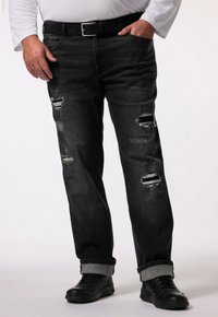 Vaqueros de mezclilla negra con rasgaduras desgastadas, con un corte recto clásico, dobladillos enrollados y un cinturón negro. Llevados con zapatos negros.