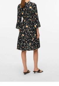 Abito nero a fiori con maniche a tre quarti, caratterizzato da fiori gialli e rosa, una vita sfrangiata e una gonna a strati. Indossato con sandali scivolosi marroni.