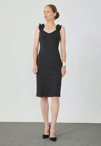 Robe noire ajustée avec un décolleté en v et des détails de manches ballon, en tissu lisse, se terminant au milieu du mollet, assortie à des escarpins noirs à bout pointu.