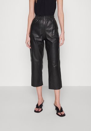 Kvinde iført sort, højtaljede, cropped læderbukser med elastisk linning og sorte åben-tå hælede sandaler mod en ensfarvet baggrund.