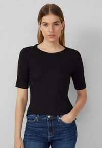 T-shirt noir à manches courtes avec un col rond, en tissu côtelé. Le mannequin l'associe à un jean en denim bleu ajusté.